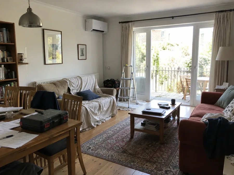 Living room on the fitting day: stepladder up, sofa under a dust sheet, unit already on the wall